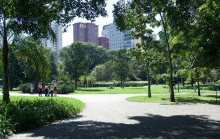 Grupa ljudi okupljena pored široke staze u zelenom gradskom parku sa zgradama u pozadini