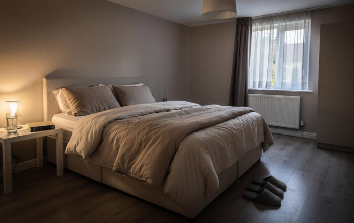 Uređen krevet sa bež posteljinom, noćnim stočićem i upaljenom lampom