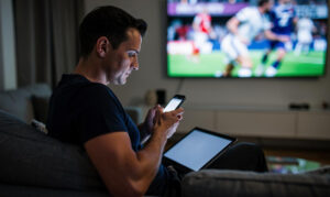 Muškarac sedi na kauču i koristi telefon dok mu tablet leži na krilu a TV u pozadini prikazuje fudbal