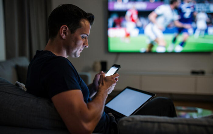 Muškarac sedi na kauču i koristi telefon dok mu tablet leži na krilu a TV u pozadini prikazuje fudbal